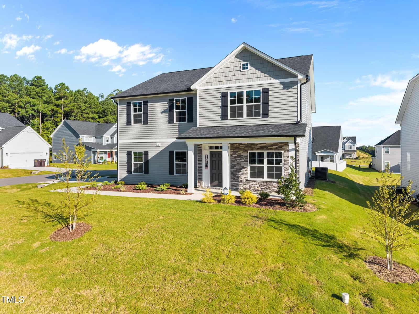 Two-story suburban house with gray siding and stone accents, black shutters, and a covered front porch. The yard is landscaped with young trees and shrubs, and neighboring homes are visible in the background under a blue sky.