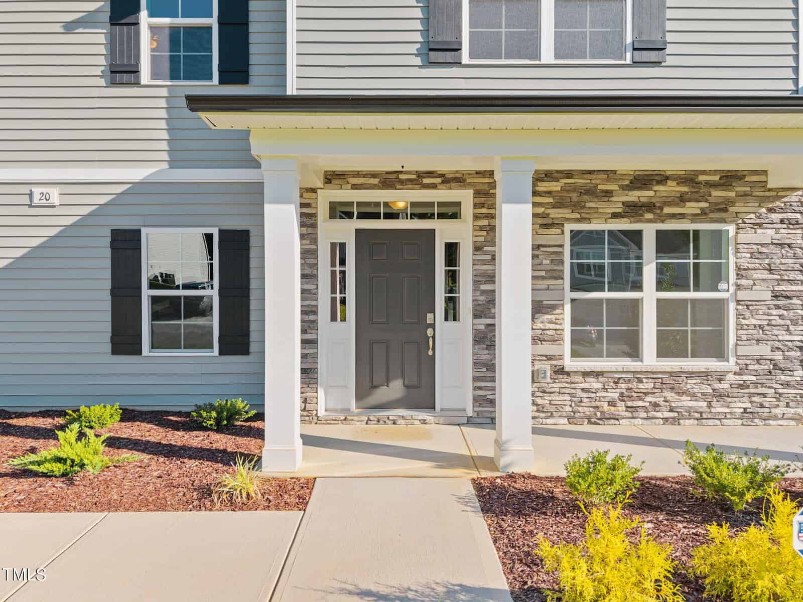 A gray front door with glass panels is centered between white columns and windows on a house exterior with gray siding and stone accents. Shrubs and mulch line the walkway leading to the entryway.