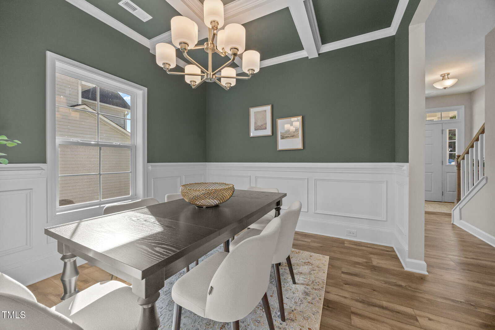 A dining room with green walls and white wainscoting, a black rectangular table with four white chairs, a gold chandelier, two framed pictures, and a window letting in natural light.