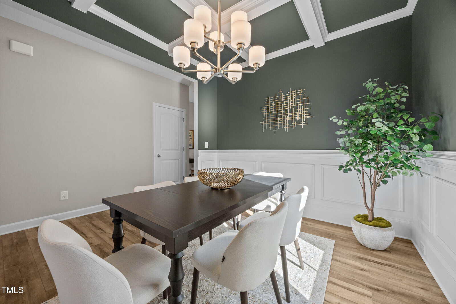 A dining room with a dark rectangular table, six white chairs, a modern chandelier, a decorative bowl, a wall sculpture, a large potted plant, wood flooring, and green and white walls with wainscoting.