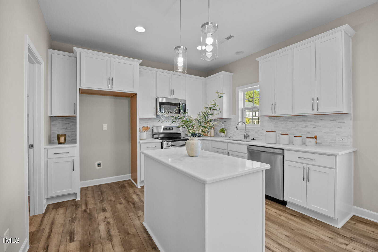 Modern kitchen with white cabinets, stainless steel appliances, a center island with a vase, wood-style flooring, and marble countertops. Pendant lights hang above the island. Light gray backsplash tiles cover the wall.