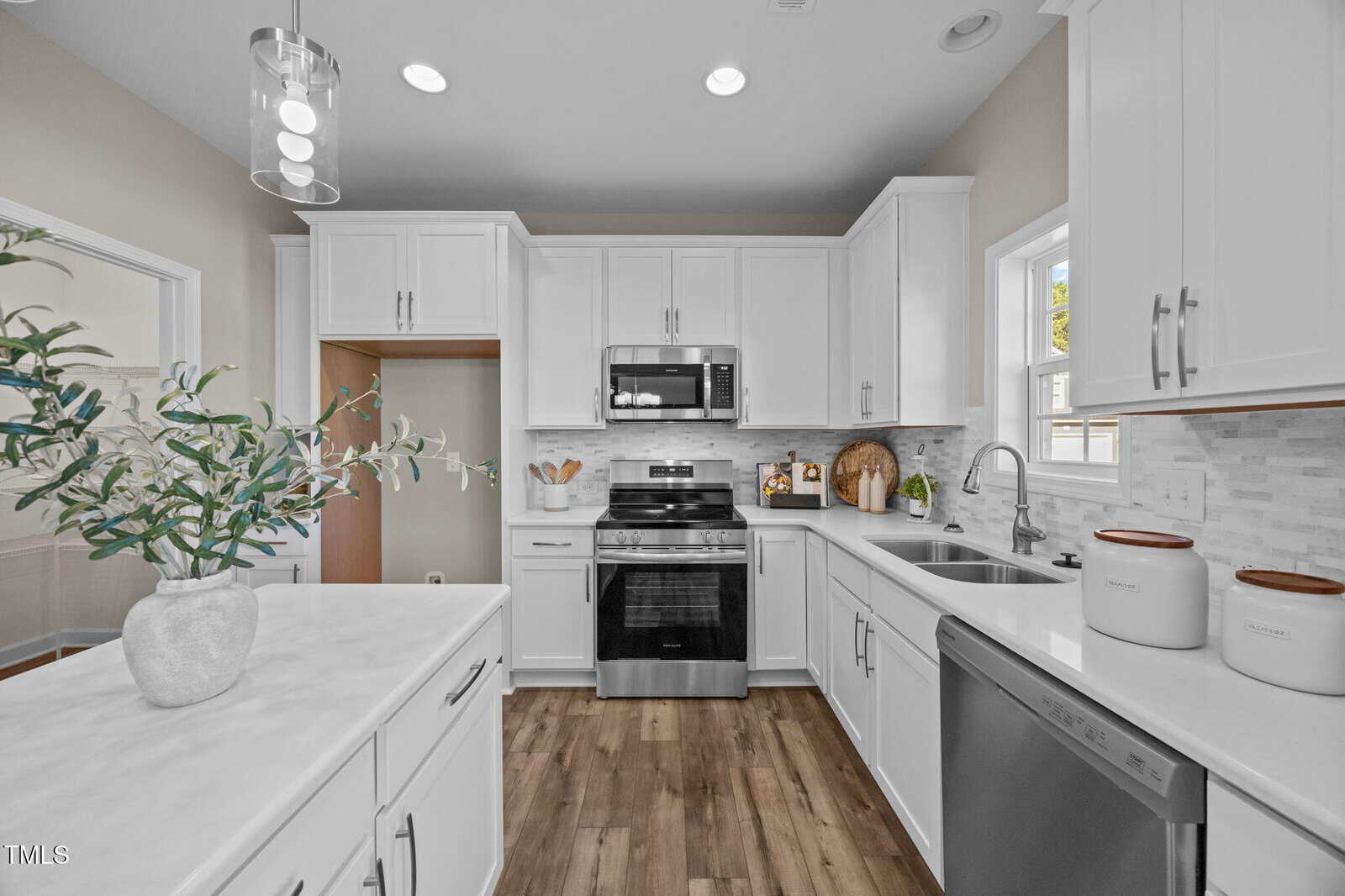 Modern kitchen with white cabinets, stainless steel appliances, a double sink, and wood flooring. A potted plant sits on the island. There are containers on the counter and a window above the sink.