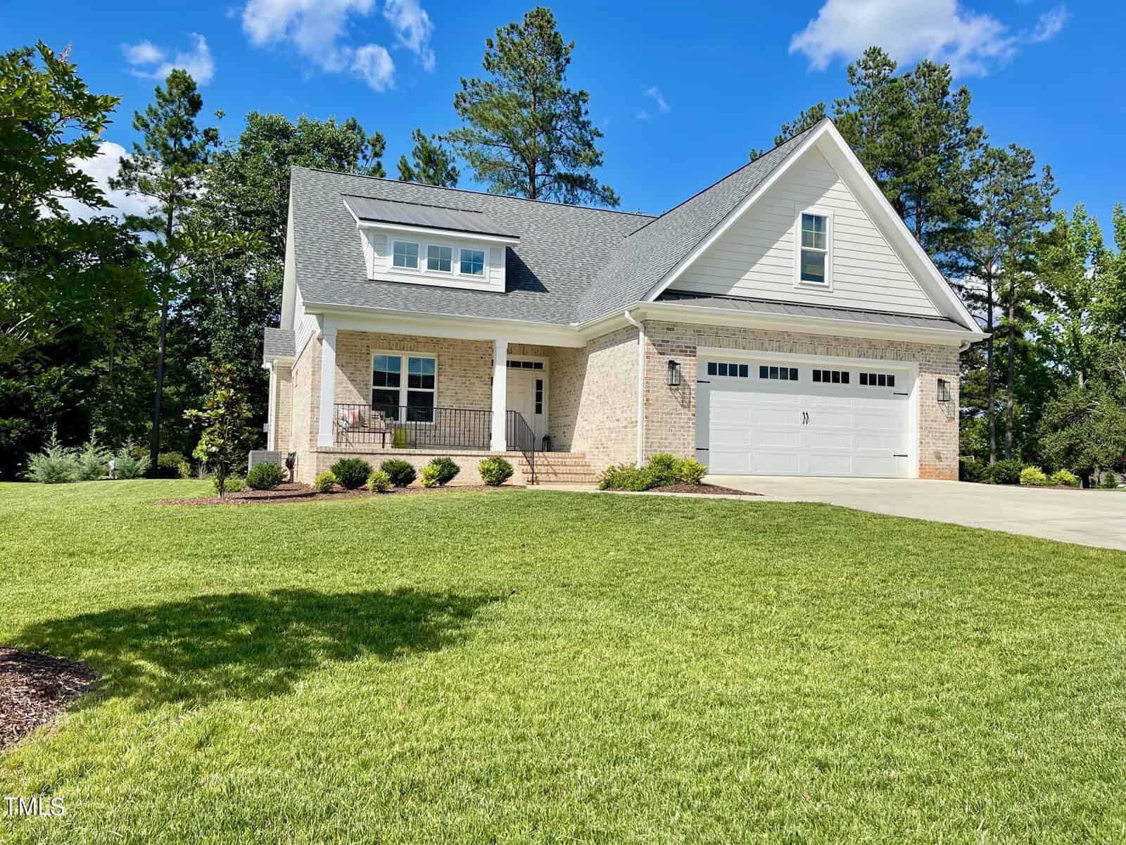 The charming two-story house at 404 Eagle Court, Henderson NC features a sloped roof, light-colored brick and siding exterior, and a front porch with black railing. With multiple windows and a two-car garage, it sits on a manicured lawn backed by tall trees under a clear blue sky.