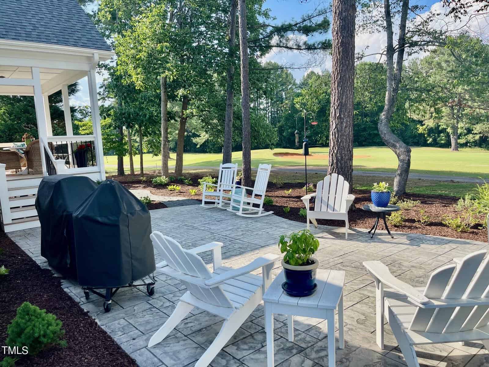 A backyard patio featuring white Adirondack chairs and tables, with potted plants and a grill. The area is surrounded by tall trees, and a manicured lawn extends to a golf course in the background. The scene is serene with a mix of sun and shade.
