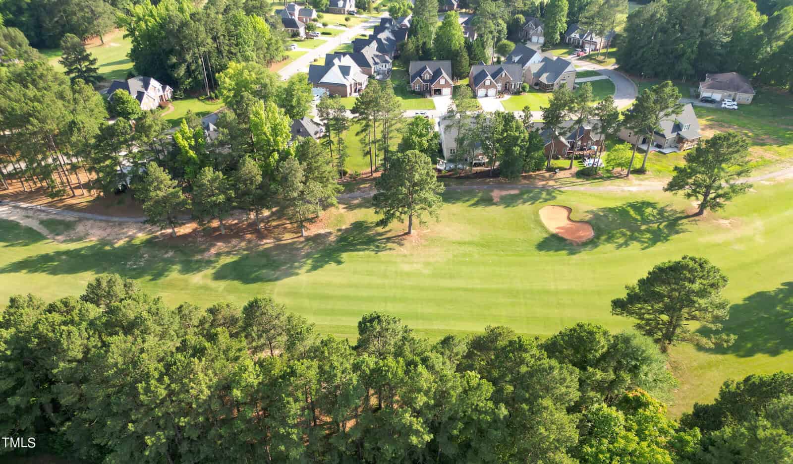 Aerial view of a suburban neighborhood with a golf course in the foreground. The area is heavily wooded around the edges, with houses featuring spacious yards and tree-lined streets, and the green expanse of the golf course is visible with a sand trap.