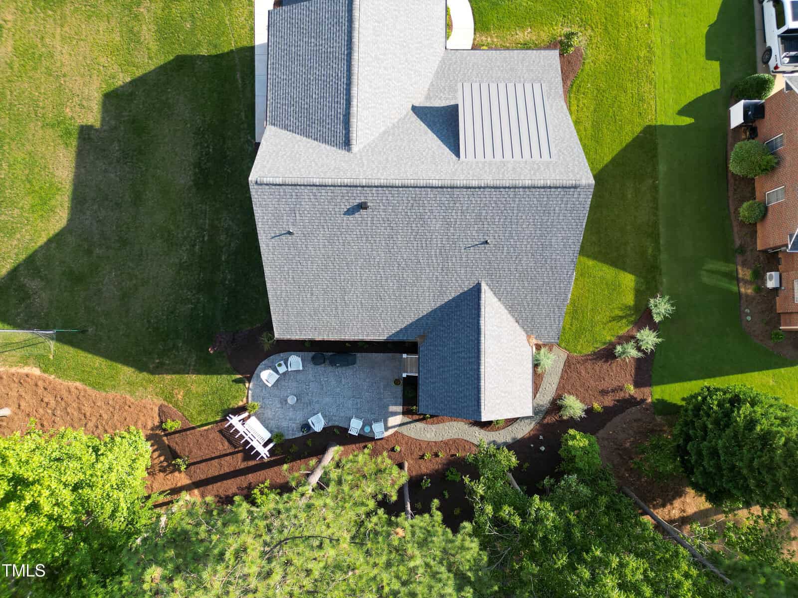 Aerial view of a house with a gray roof, a patio with outdoor furniture, and surrounding landscaped garden. The patio area features a circular table with chairs and a white lounge chair. Lawn and trees are visible in the well-maintained garden.