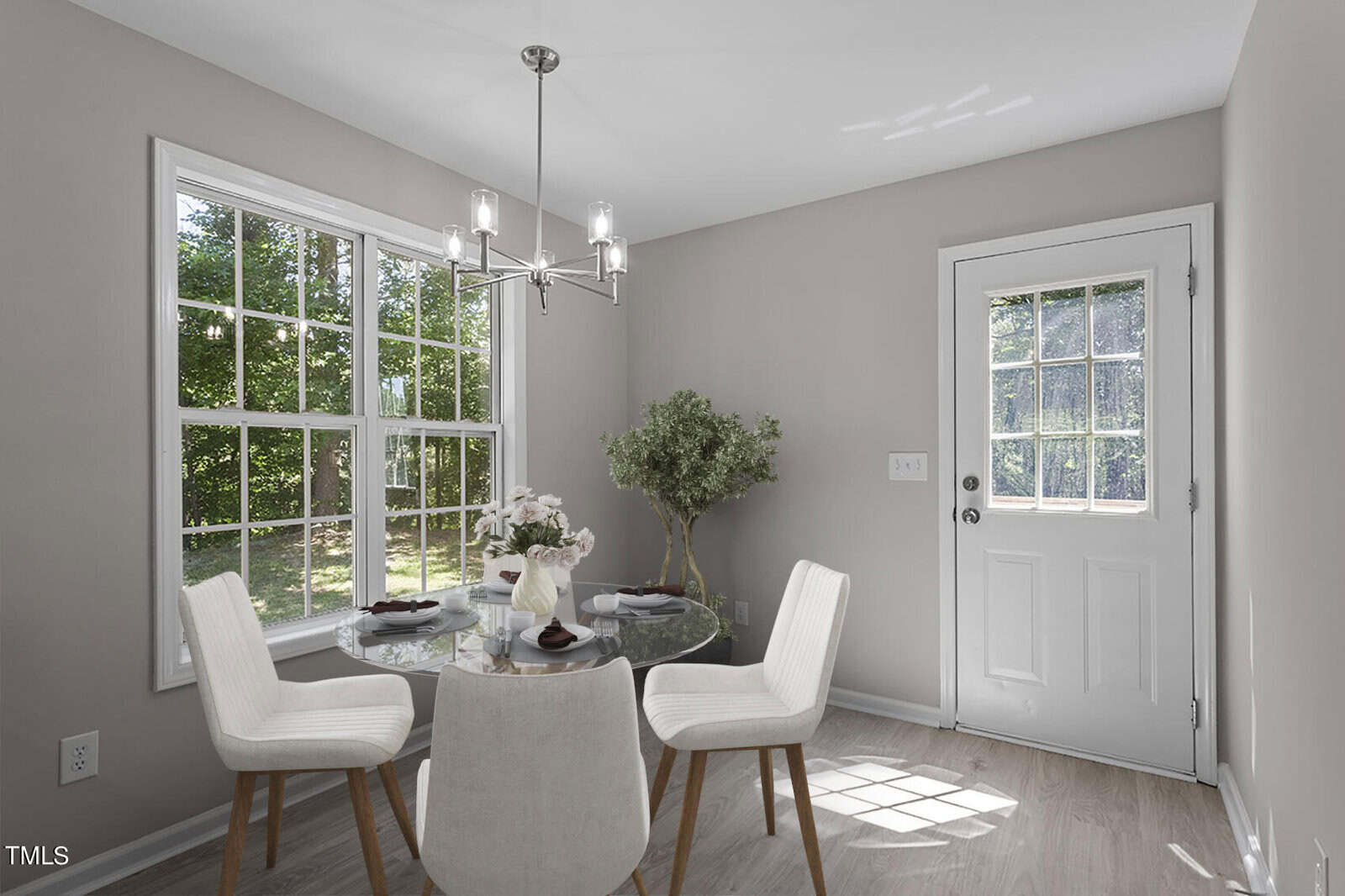 A small dining area with a round glass table, set with white plates and a plant centerpiece. Three white chairs surround the table. Large windows and a glass-paneled door let in natural light. A chandelier hangs above, and a potted plant is in the corner.