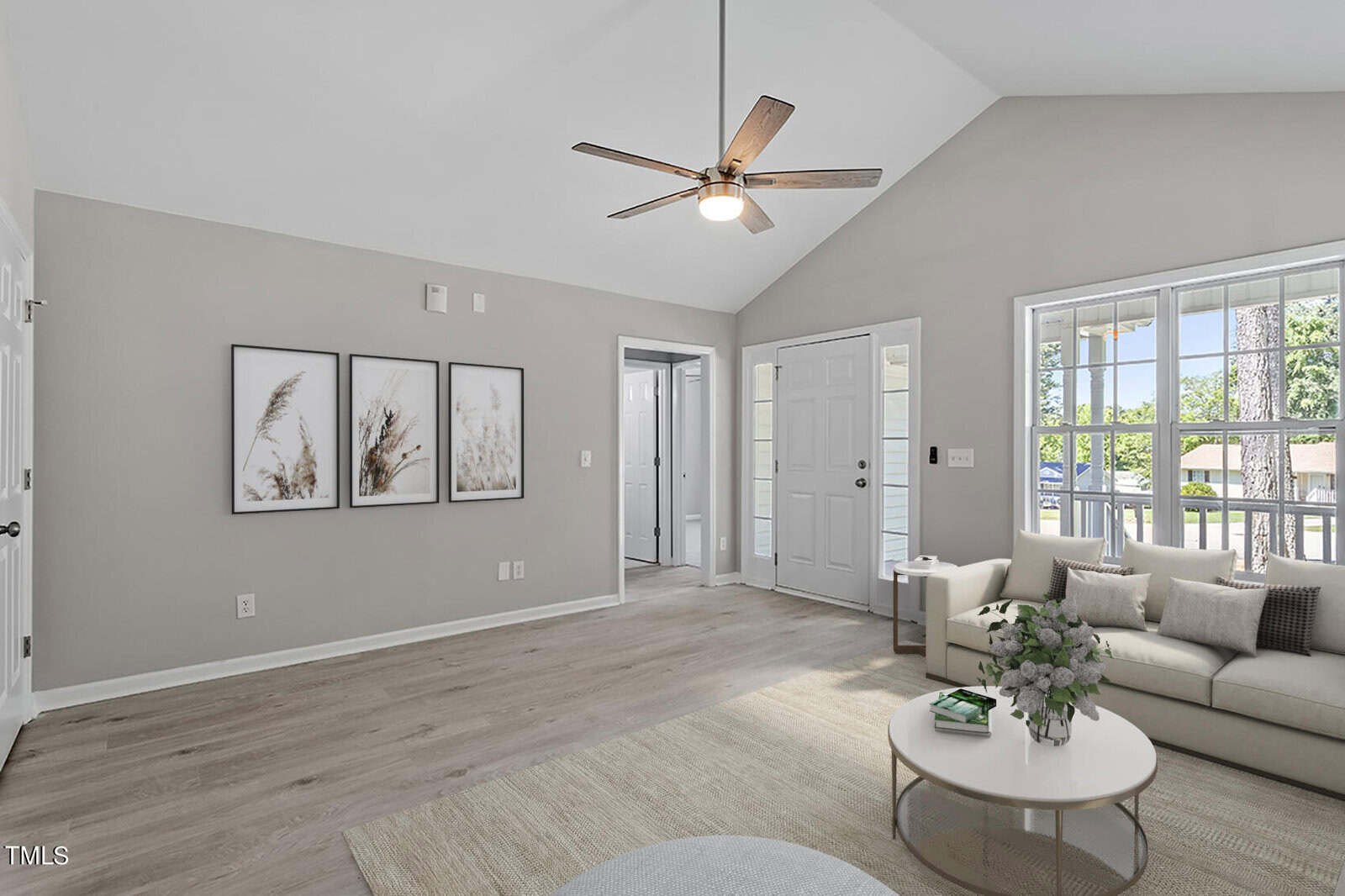 Spacious living room with light gray walls and wooden flooring. A modern ceiling fan hangs above. The room features a gray sofa, a round coffee table, and three nature-themed framed artworks on the wall. Large windows allow natural light in.