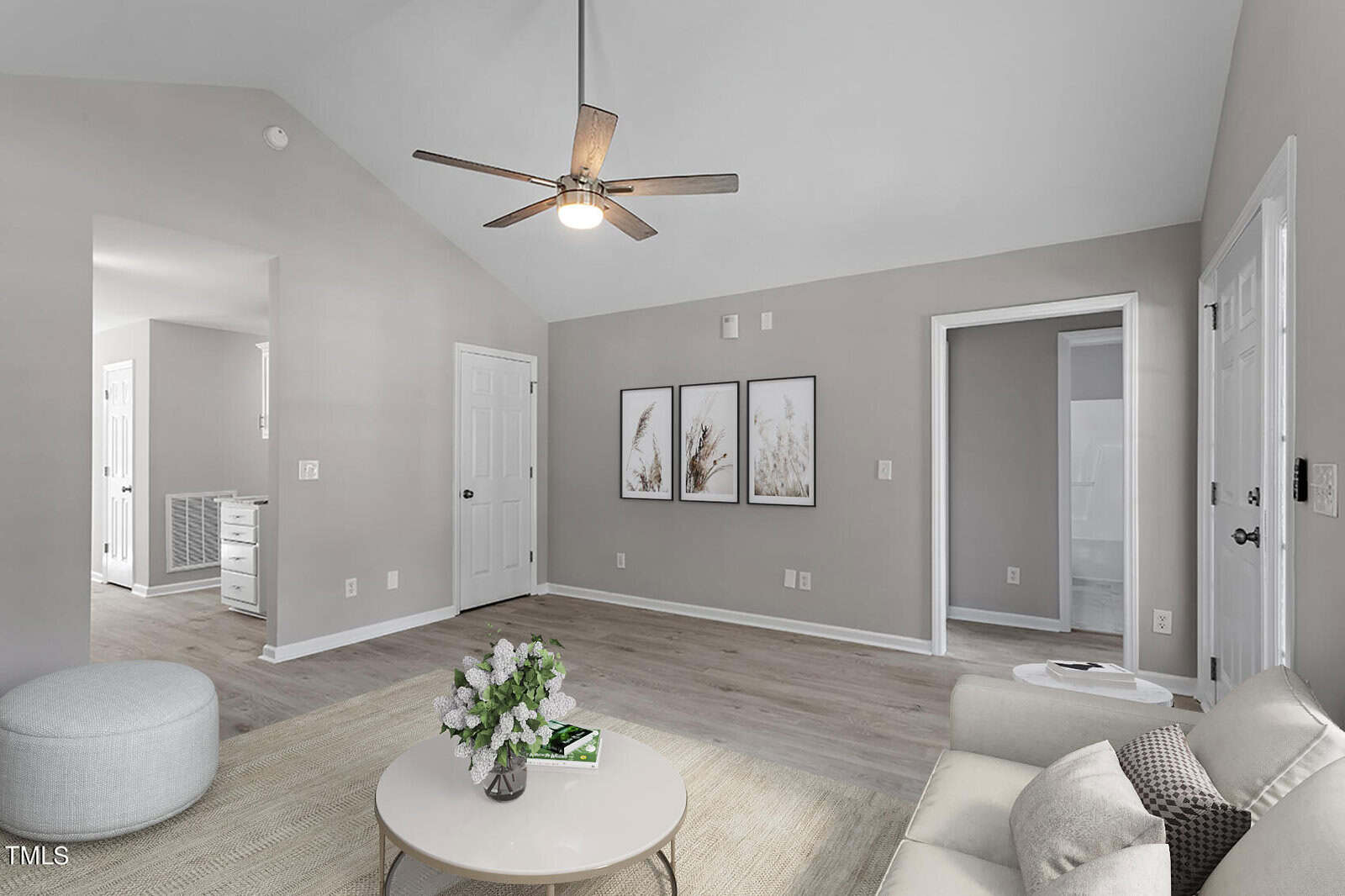 A modern living room with gray walls and light wood flooring. It features a beige sofa, a round coffee table with flowers, and three framed artworks on the wall. There's a ceiling fan and a glimpse of an adjacent hallway.
