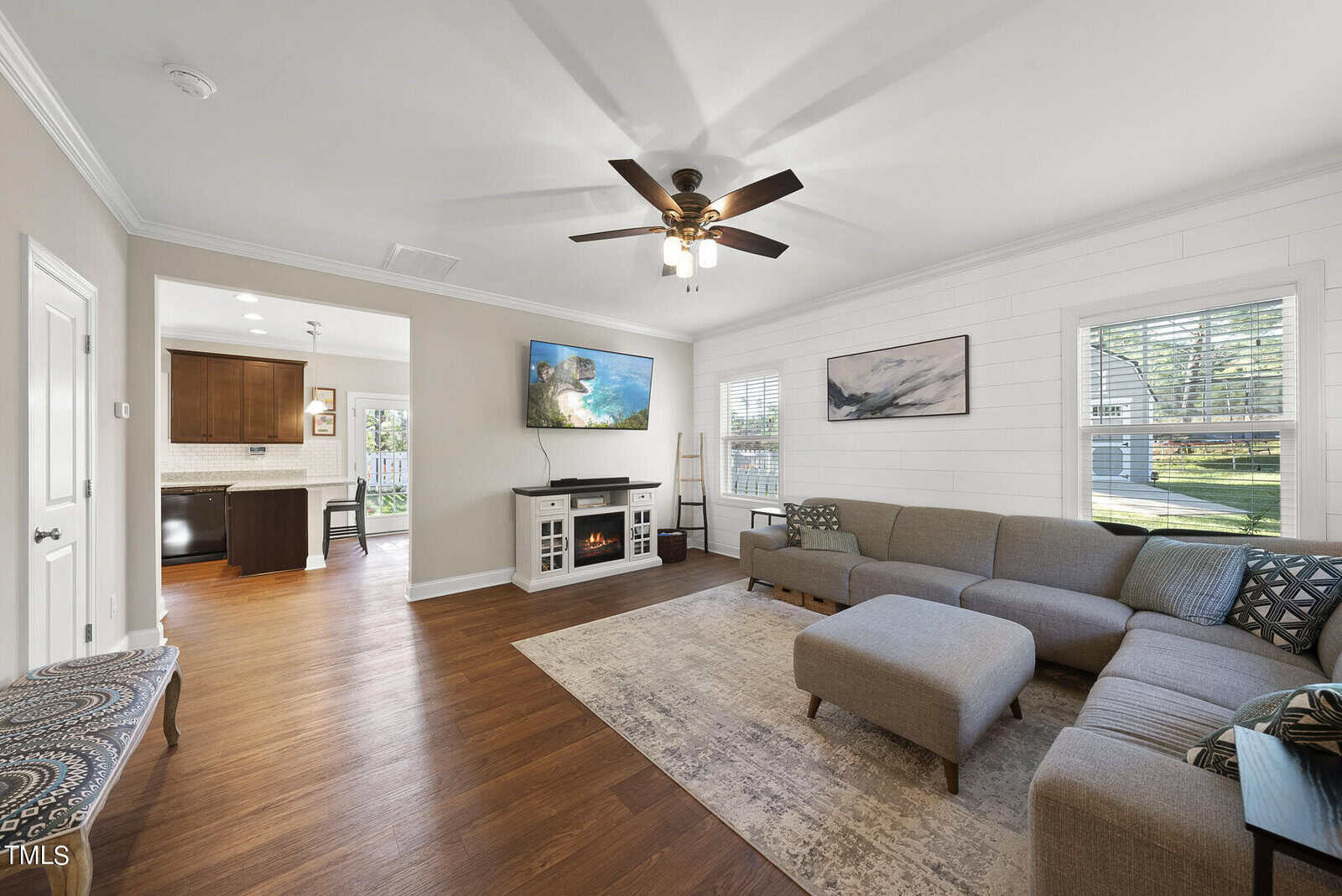 A cozy living room with a large gray sectional sofa, a ceiling fan, and a wall-mounted TV. The room has wooden floors and white walls with a shiplap accent. A window with blinds lets in natural light. The adjacent kitchen is visible in the background.