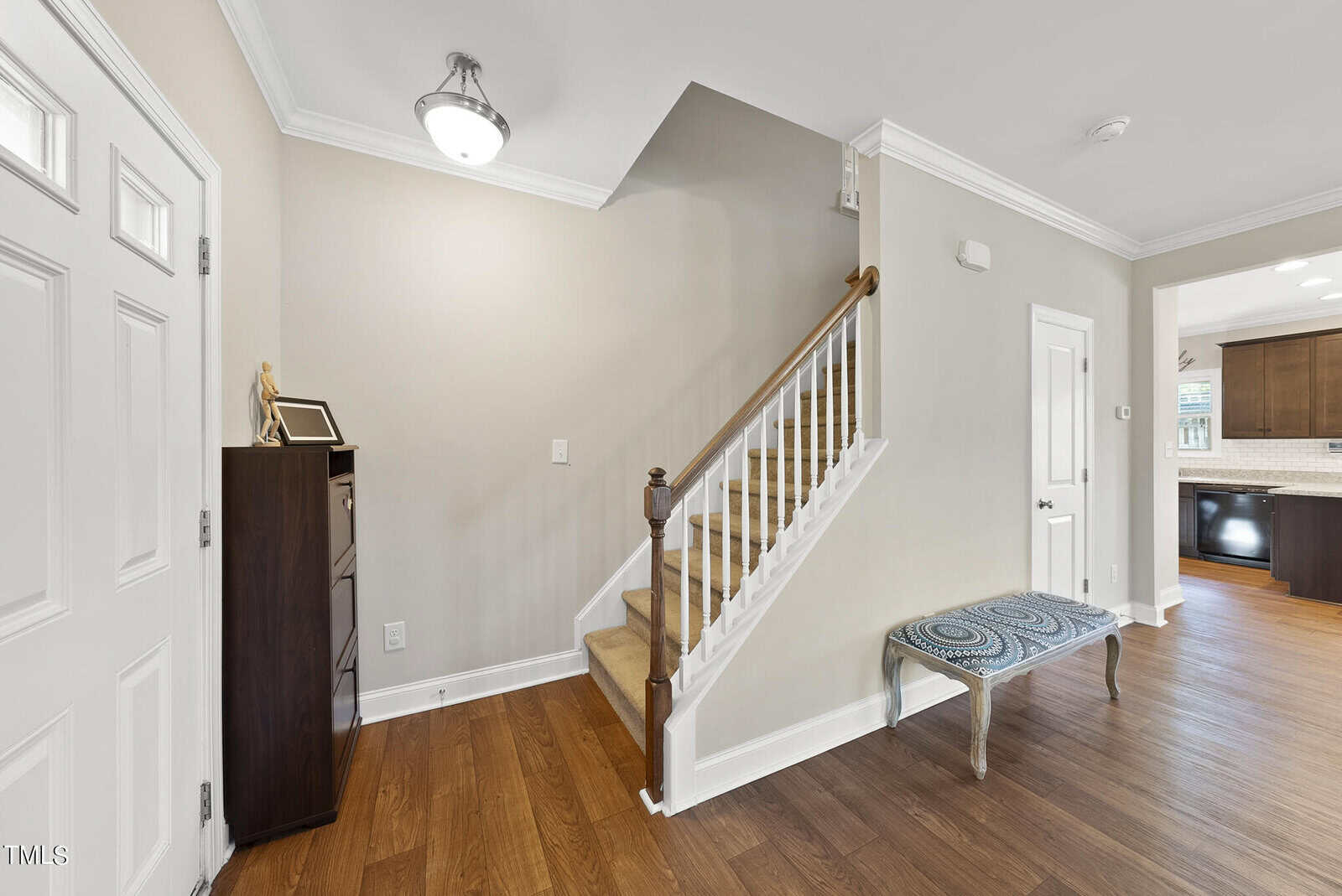 A hallway with a wooden staircase, beige walls, and a small decorative table. A closed door is on the left, leading to an open kitchen area. A framed picture rests on a dark cabinet beneath a ceiling light.