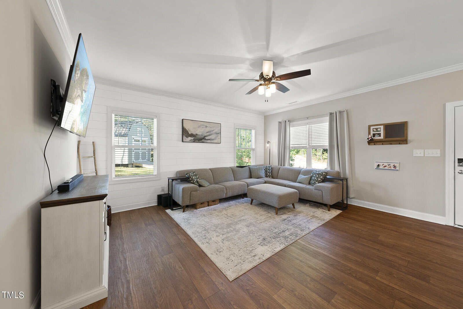 Bright living room with a gray sectional sofa, a wall-mounted TV, and wooden floors. The room features large windows, a ceiling fan, a light gray rug, and minimal decor, creating a modern and cozy atmosphere.