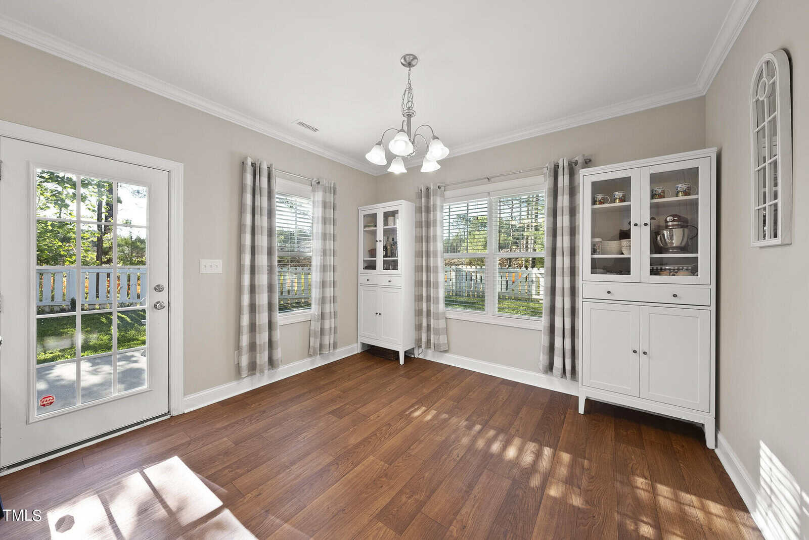 A bright dining room with wooden flooring, featuring two white cabinets, plaid curtains on large windows, and a glass-door entry leading outside. A modern chandelier hangs from the ceiling, illuminating the space.