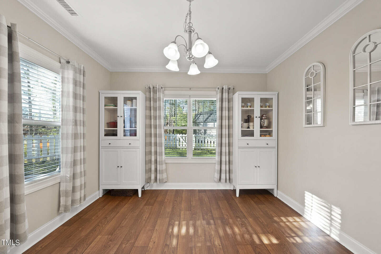 Bright dining room with wooden floor, two white cabinets with glass doors, and a window with patterned curtains. A chandelier hangs from the ceiling, and two decorative mirrors are on the right wall.