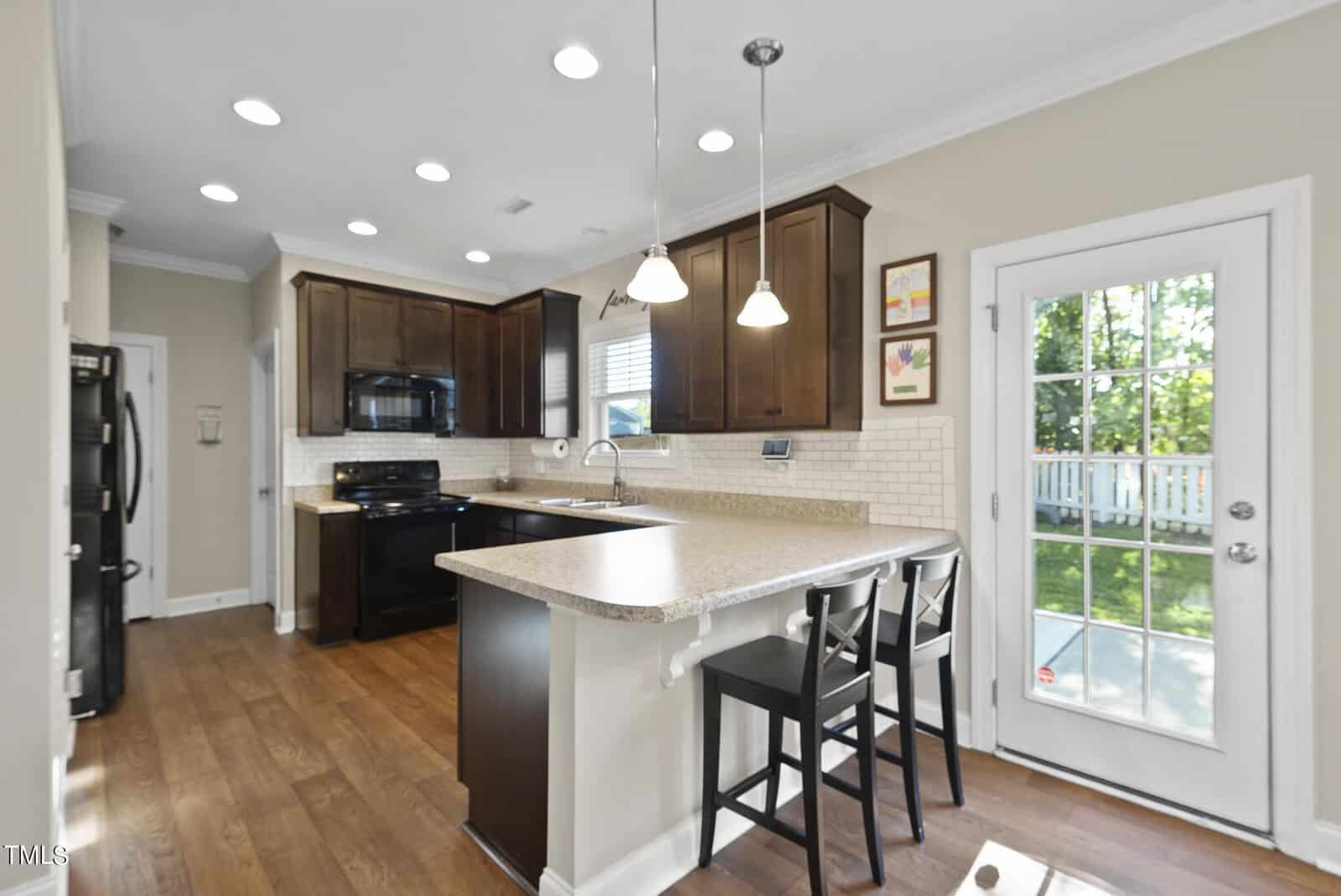 A bright kitchen with dark wood cabinets, granite countertops, and an island with two chairs. Pendant lights hang above the island. There's a window and a door leading to a garden with sunlight filtering through.