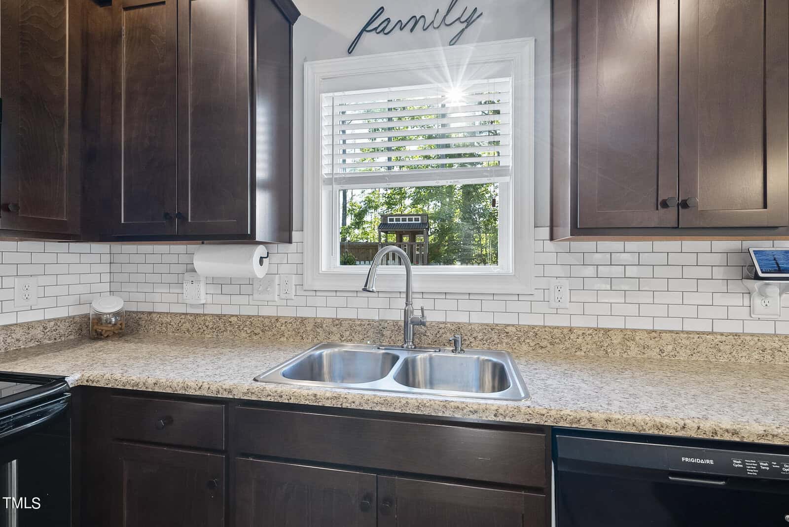 A kitchen sink with a double basin is centered under a window with sunlight streaming in. The countertop is beige with a speckled pattern, and dark wooden cabinets surround it. A paper towel holder and a small jar are positioned nearby.