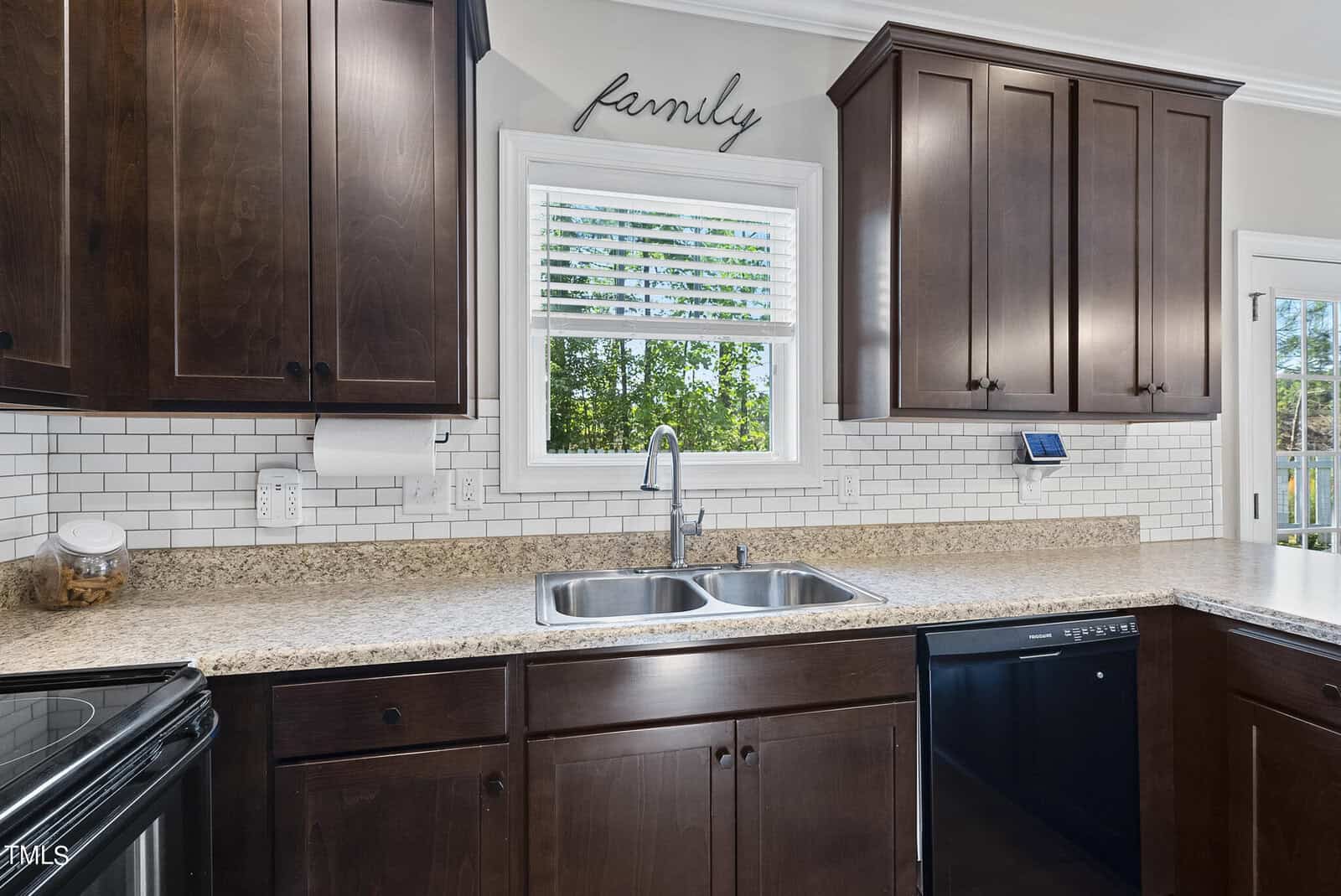 A kitchen with dark wood cabinets, a double sink, and granite countertops. There's a window above the sink with a view of trees. The word "family" decorates the wall above the window, and appliances are visible on the counter.