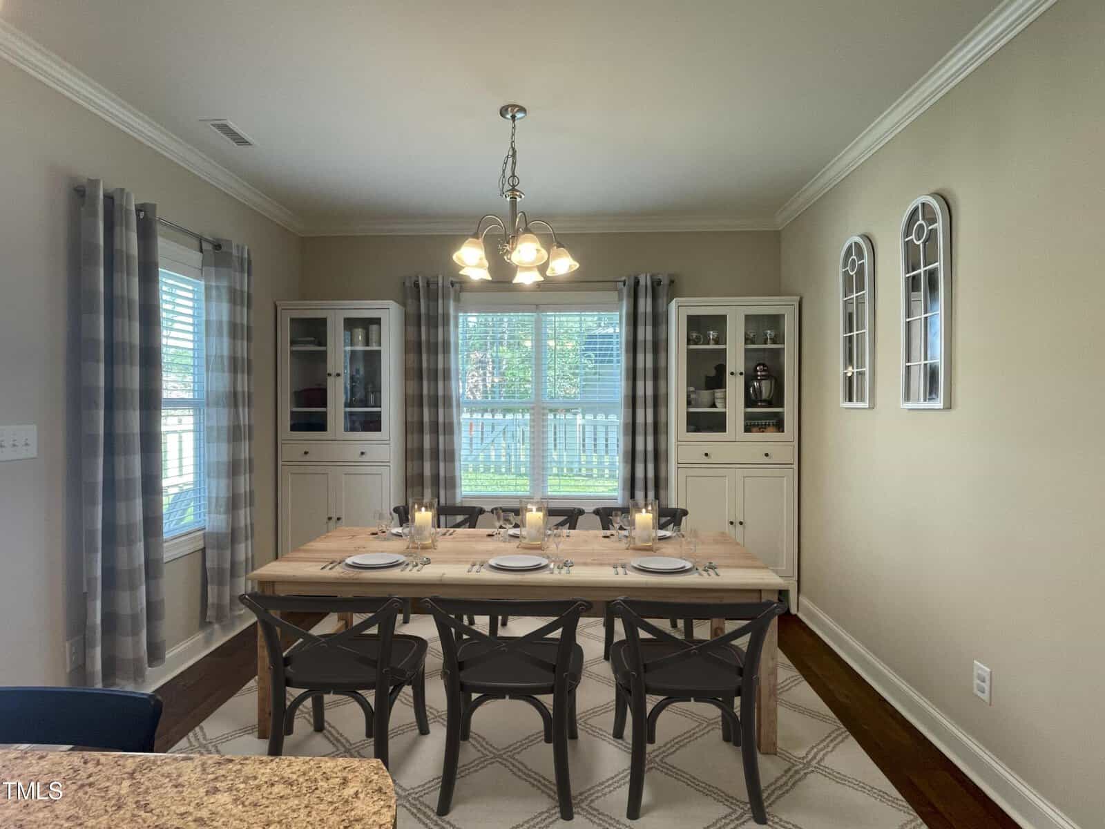A dining room with a wooden table set for eight, featuring candles and plates. Two glass-front cabinets flank a large window with plaid curtains. A chandelier hangs above, and two decorative mirrors adorn the wall. The room has light-colored walls and flooring.