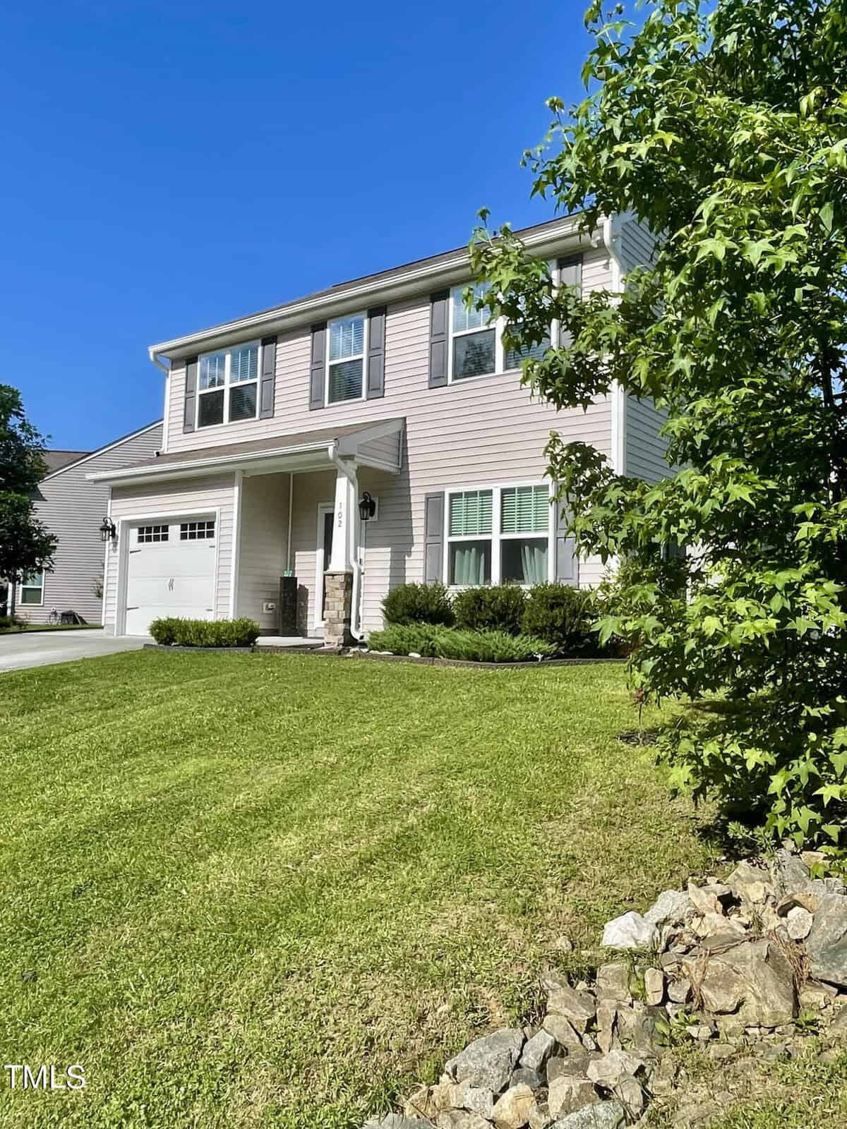 Nestled at 102 Richmond Run, this two-story house features light beige siding and a single garage door next to a charming small front porch. A neatly trimmed green lawn and a tall tree on the right enhance its appeal, all set under a clear blue sky.