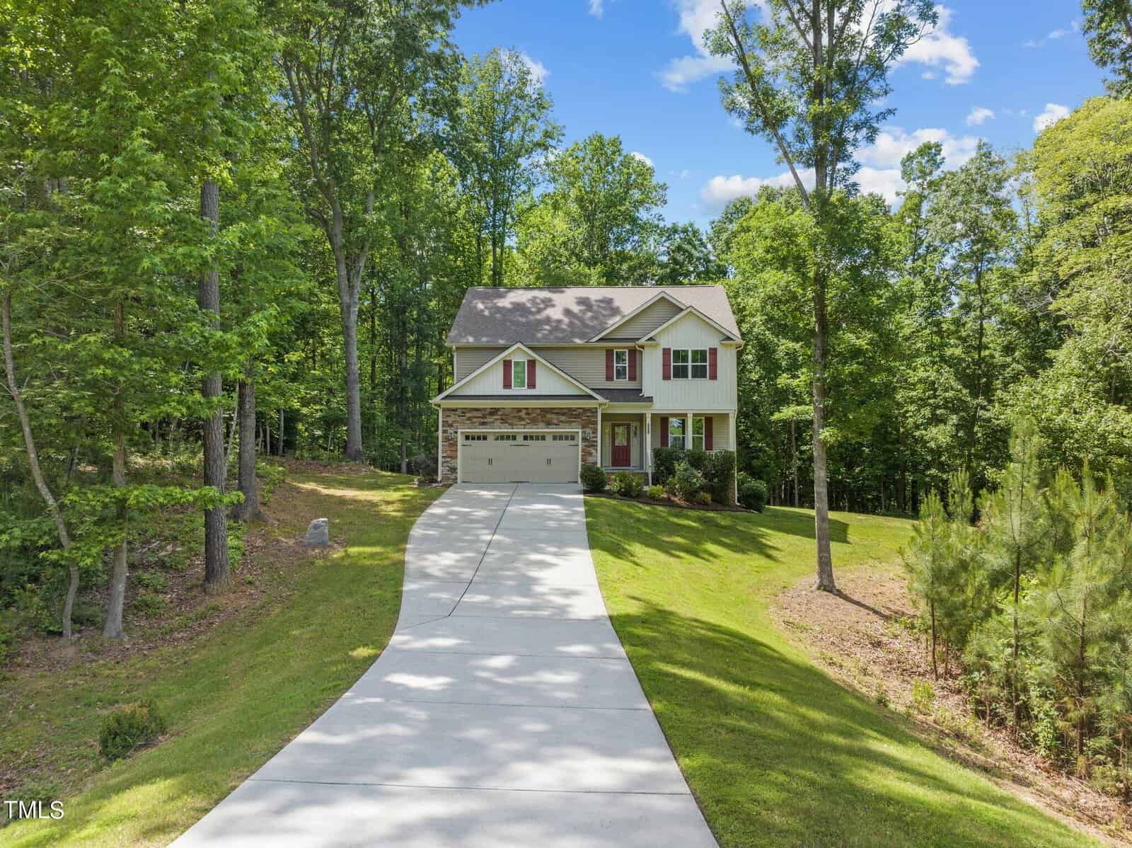 Nestled in the serene surroundings of 1321 Sourwood Drive, this two-story house features a beige exterior with charming red shutters. Located in Wake Forest, it boasts a long, winding driveway leading to an attached two-car garage, all embraced by well-maintained, lush green lawns and towering trees.