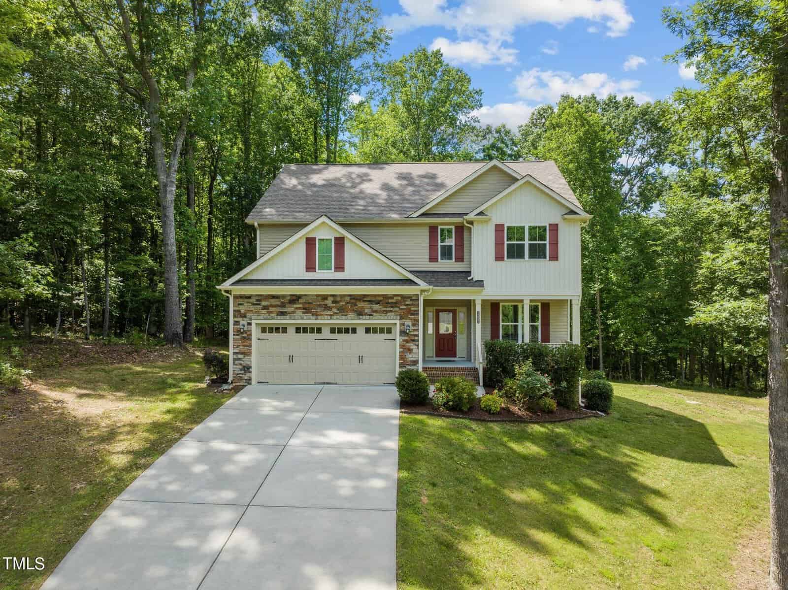 A beige two-story house with a gray roof, red shutters, and a garage nestled among lush trees. The front yard features a concrete driveway and neatly trimmed grass. The background shows a dense forest and a blue sky with scattered clouds.