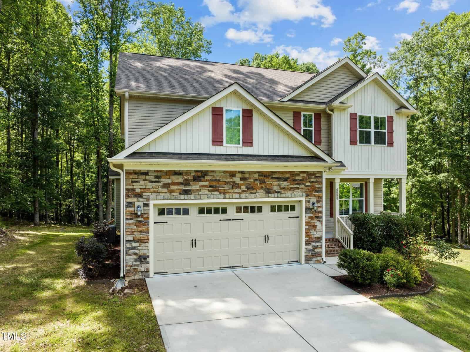 A two-story house with a brick and siding exterior, featuring a double garage door. It is surrounded by a lush, wooded area with a paved driveway leading to the entrance. The sky is clear with a few clouds.