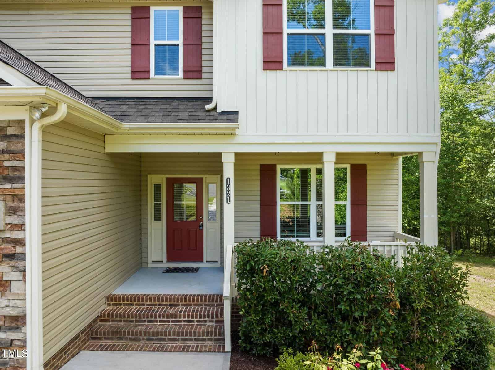 A two-story house with beige siding and red shutters. There is a porch with a white railing and brick steps leading to a red front door. Bushes line the front of the porch, and trees are visible in the background.