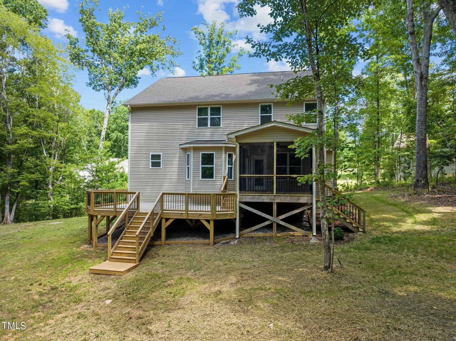Two-story house with beige siding and several windows is set against a backdrop of trees. The back features a spacious wooden deck with stairs leading down to a grassy yard. A covered, screened-in porch is visible on the right side of the deck.