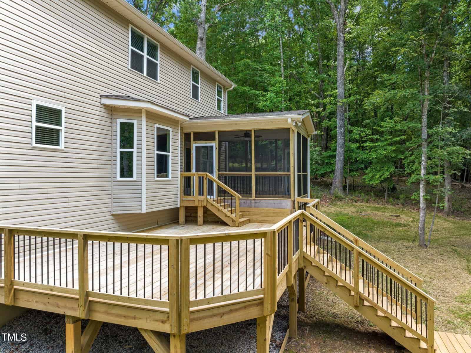 A two-story tan house with a large wooden deck and stairs leading to the backyard. The deck features a screened-in porch. The house is surrounded by lush green trees.