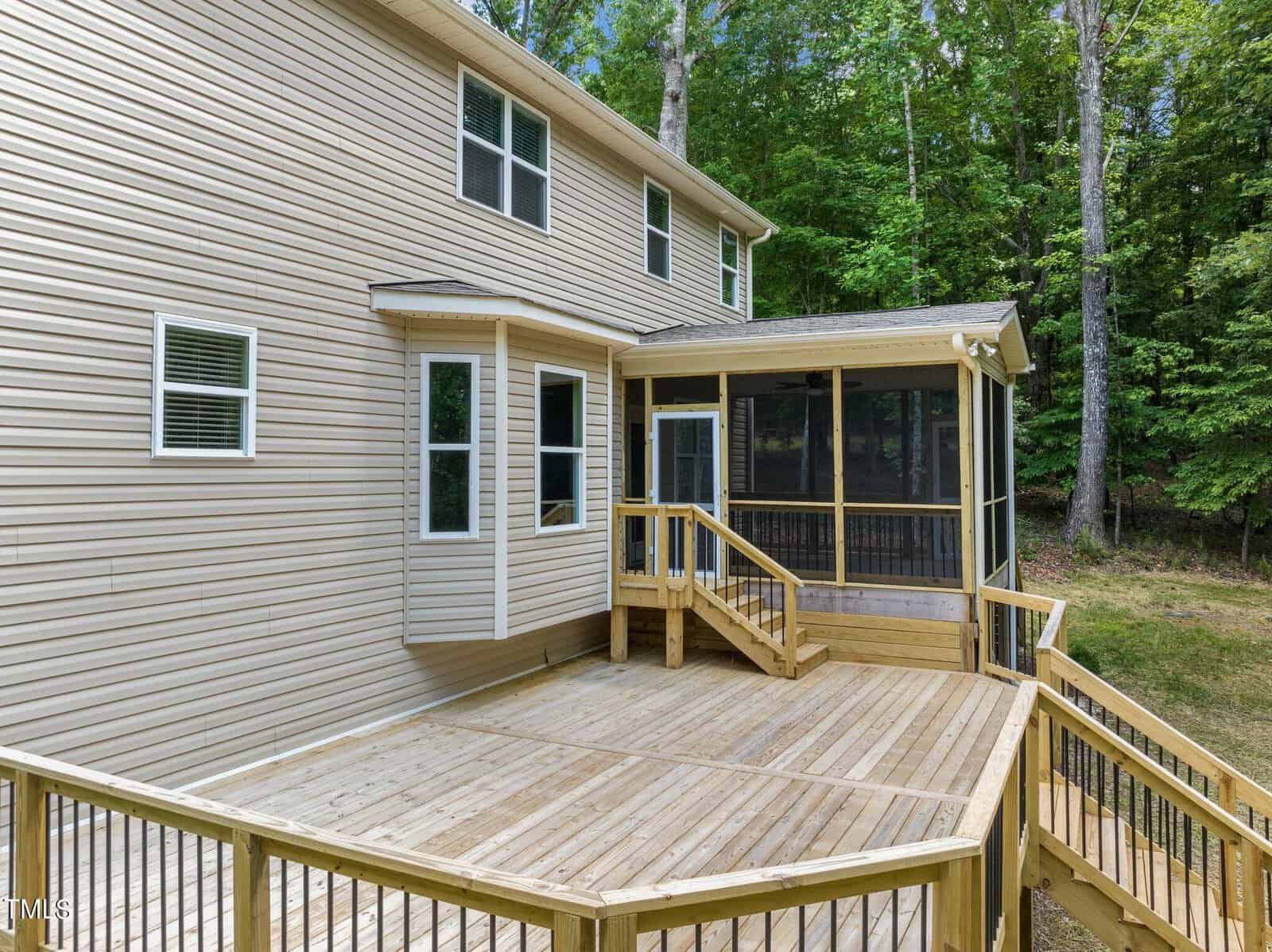A two-story beige house with a spacious wooden deck and screened porch. Surrounded by a wooded area, the deck features steps leading down and glass windows on the house's rear facade.