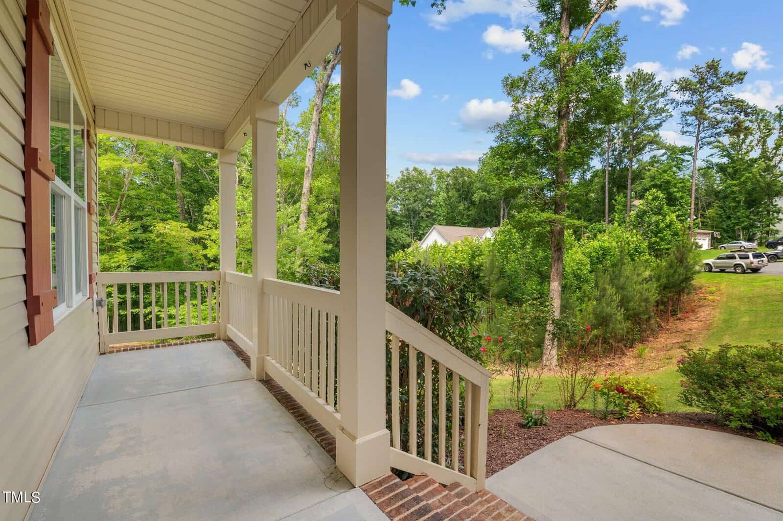 A view from a porch with white railings overlooking a lush green garden with trees and shrubs under a clear blue sky. The concrete path leads to the front yard, where a house is partially visible in the background.