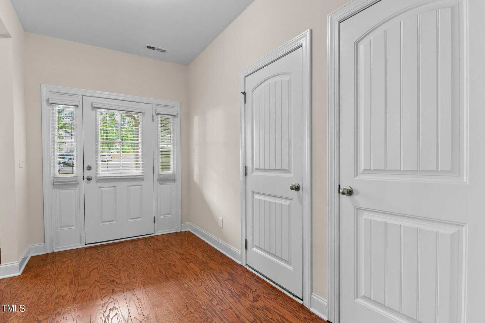 A hallway with wooden floors, two white paneled doors on the right, and a double glass door with side windows leading outside. Sunlight streams through the glass, creating a bright and inviting atmosphere.