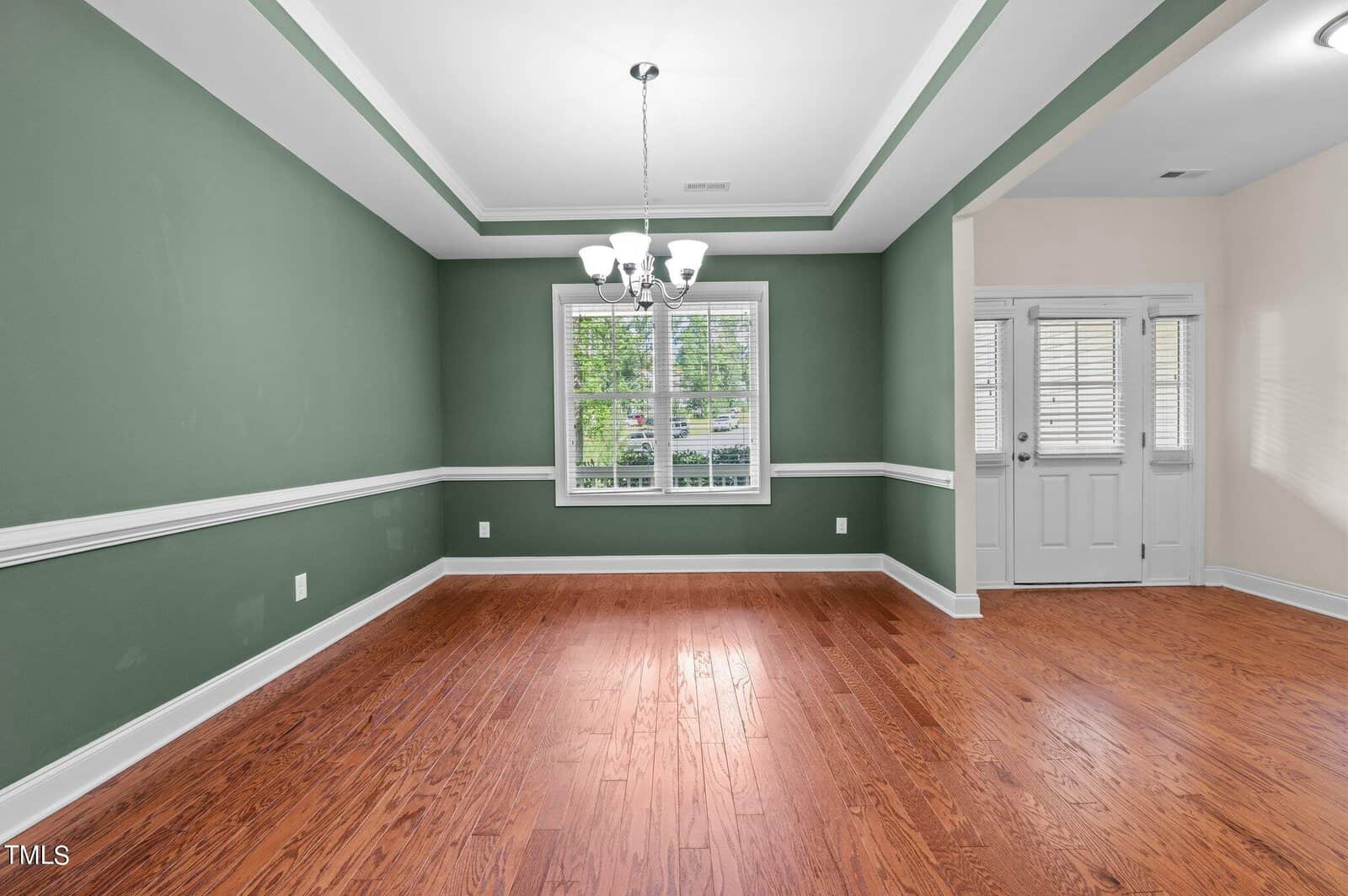An empty dining room with green walls and white wainscoting. It features a ceiling light fixture and a window with white blinds, letting in natural light. The floor is wooden, and an entryway with double doors is visible on the right.