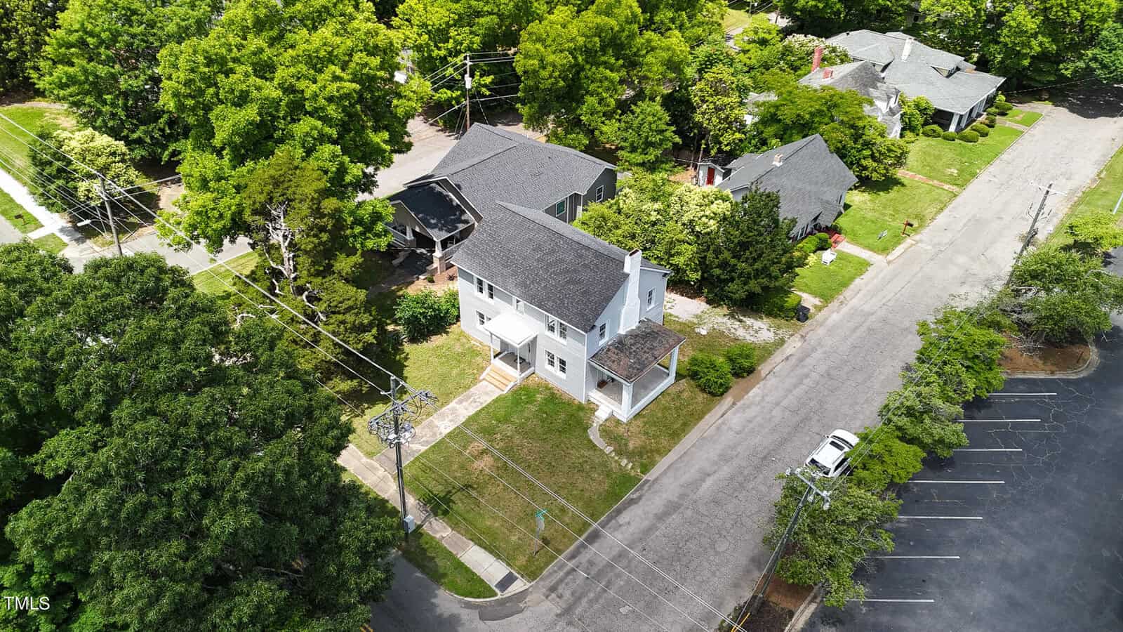 Aerial view of a residential neighborhood with tree-lined streets. The image features several houses, including a white two-story house at the center with a shaded yard. Adjacent streets and part of a parking lot are visible with parked cars and surrounding greenery.
