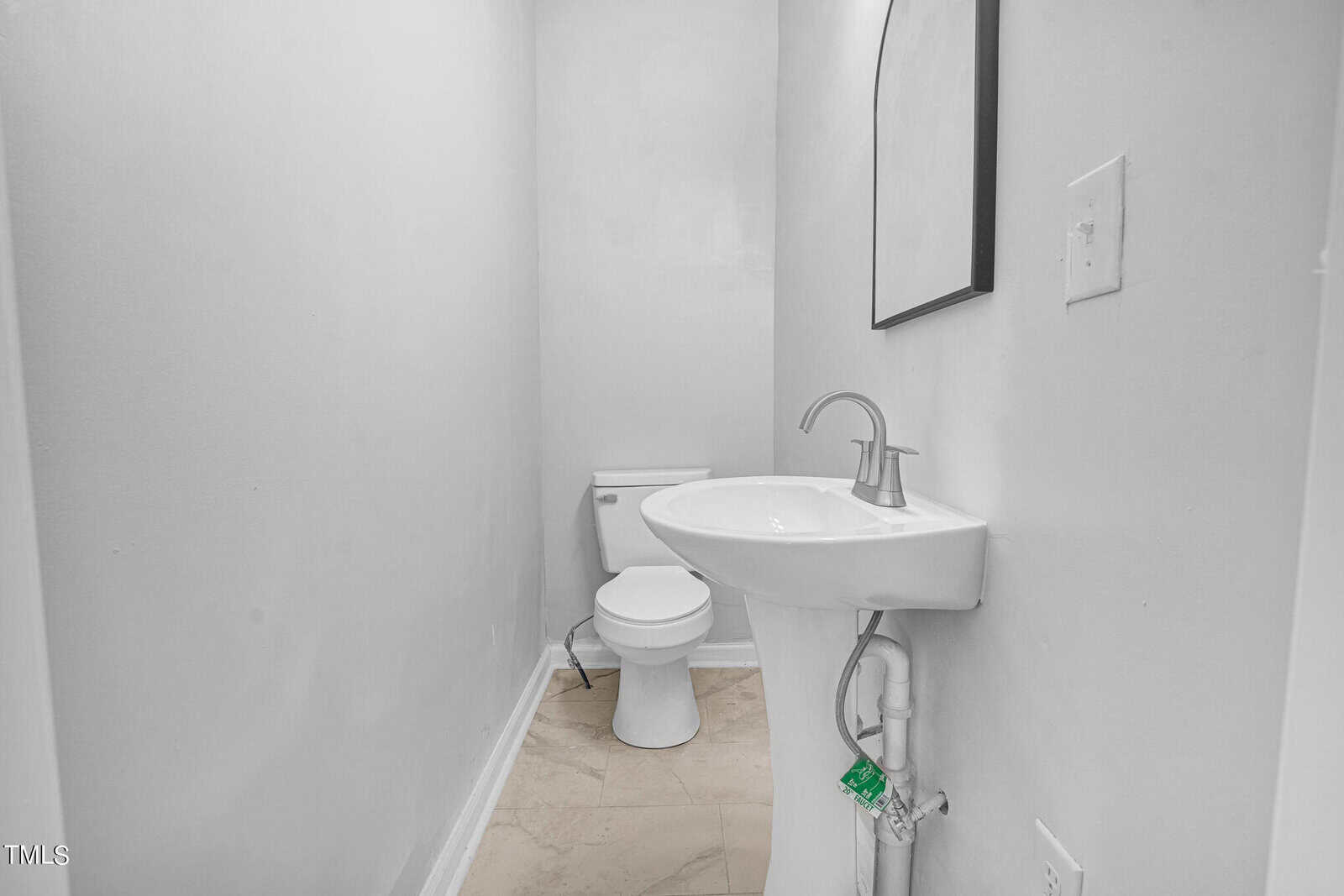 A narrow bathroom with white walls features a pedestal sink with a silver faucet, a rectangular mirror above the sink, and a white toilet against the back wall. The floor is tiled with light-colored rectangular tiles.