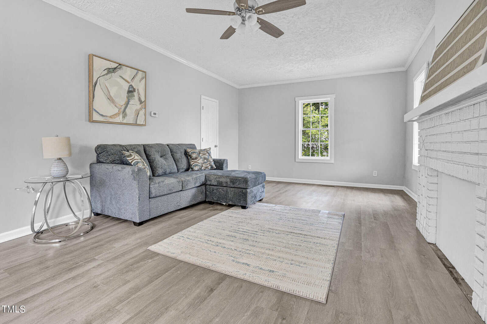 A bright, empty living room with light gray walls and hardwood flooring. A gray sectional sofa with patterned cushions sits on a rug. There's a side table with a lamp next to the sofa, a ceiling fan, a window, and a brick fireplace on the right wall.