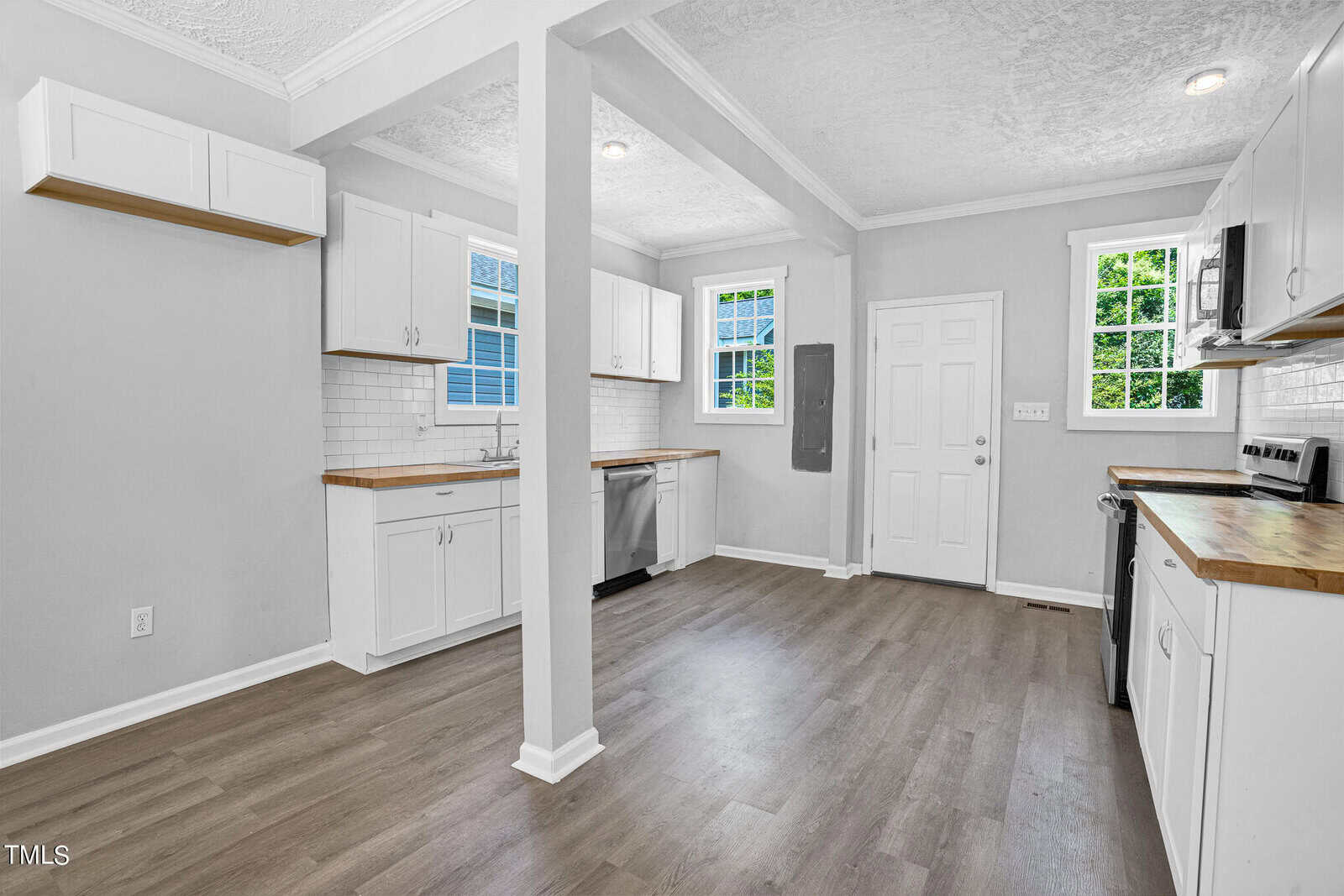 A modern kitchen featuring white cabinets, butcher block countertops, and stainless steel appliances including a stove and dishwasher. The room has light wood flooring, white subway tile backsplash, a chalkboard on the wall, and large windows providing natural light.