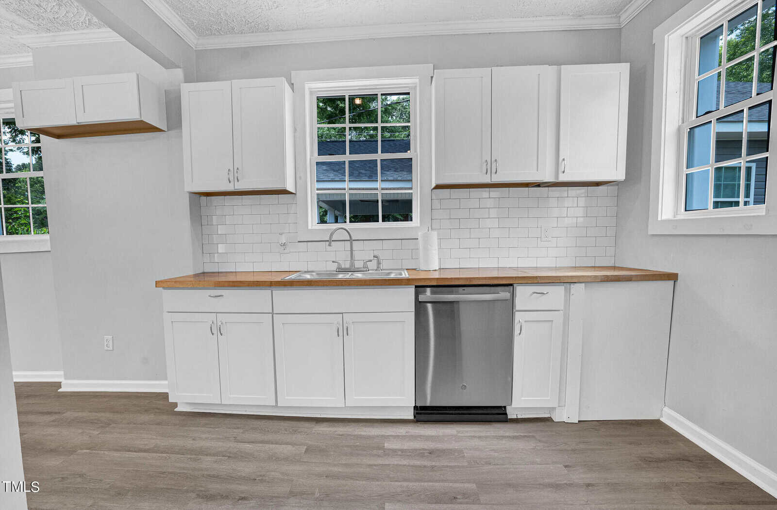 A modern kitchen with white cabinets, wood countertops, and a white subway tile backsplash. There is a stainless steel sink under the window, a built-in dishwasher to the right, and wood flooring. Large windows bring in natural light, brightening the space.