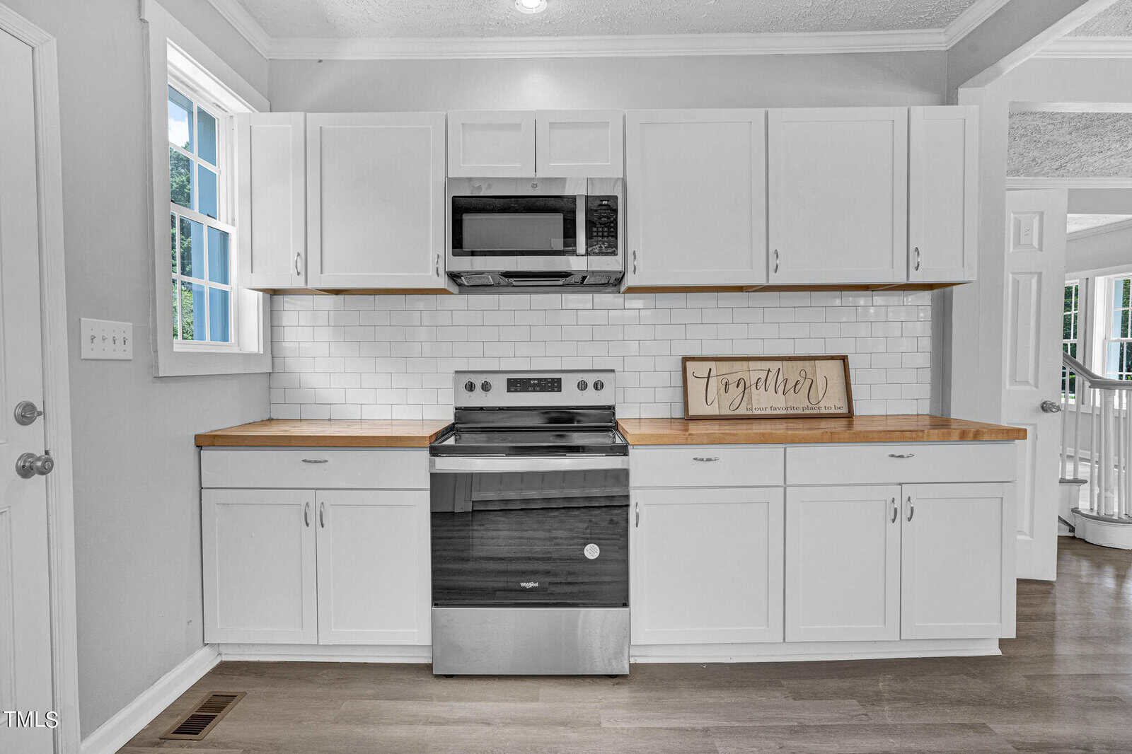 A modern kitchen features white cabinets with silver handles, light wood countertops, a white subway tile backsplash, and stainless steel appliances, including a stove with an overhead microwave. A framed sign that says "together" rests on the right counter.
