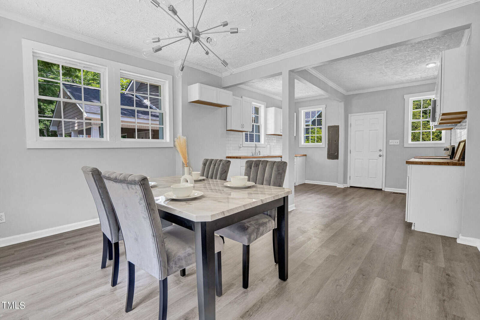 A bright, modern dining area with a rectangular table set for four. Grey upholstered chairs surround the table, which holds a simple centerpiece and white dishware. The room has large windows, light-colored walls, and a contemporary chandelier hanging from the ceiling.