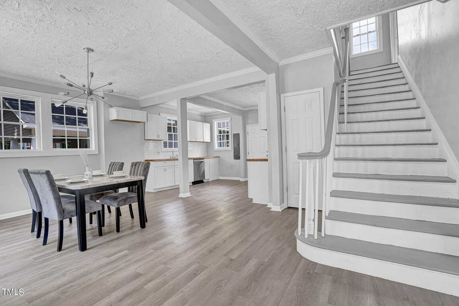 A modern dining area with gray upholstered chairs and a rectangular table set for a meal. The dining space opens into a white kitchen with upper and lower cabinets and stainless steel appliances. A wooden staircase with white railings leads to the upper floor.