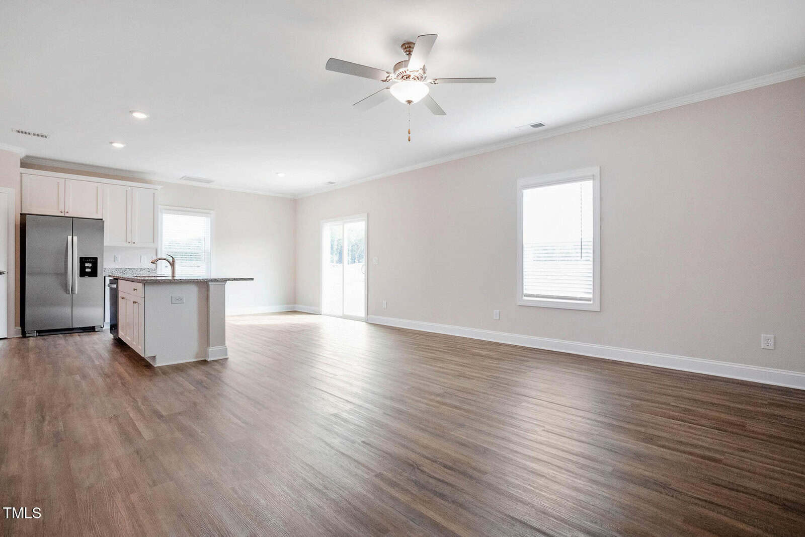 Spacious empty living room with light wood flooring and neutral walls. A ceiling fan is centered above. The room opens to a kitchen area featuring a large island, white cabinetry, and stainless steel appliances. Windows allow natural light to enter.