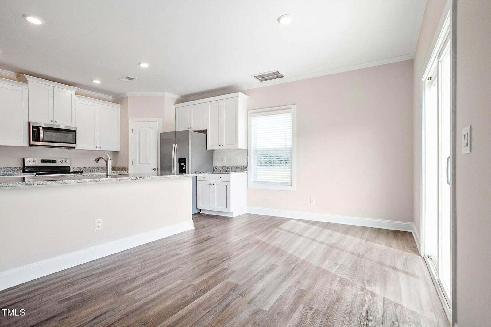 A spacious kitchen and dining area with light-colored walls and wood flooring. The kitchen has white cabinets, a granite countertop, stainless steel appliances, and recessed lighting. A sliding glass door and a window provide natural light.