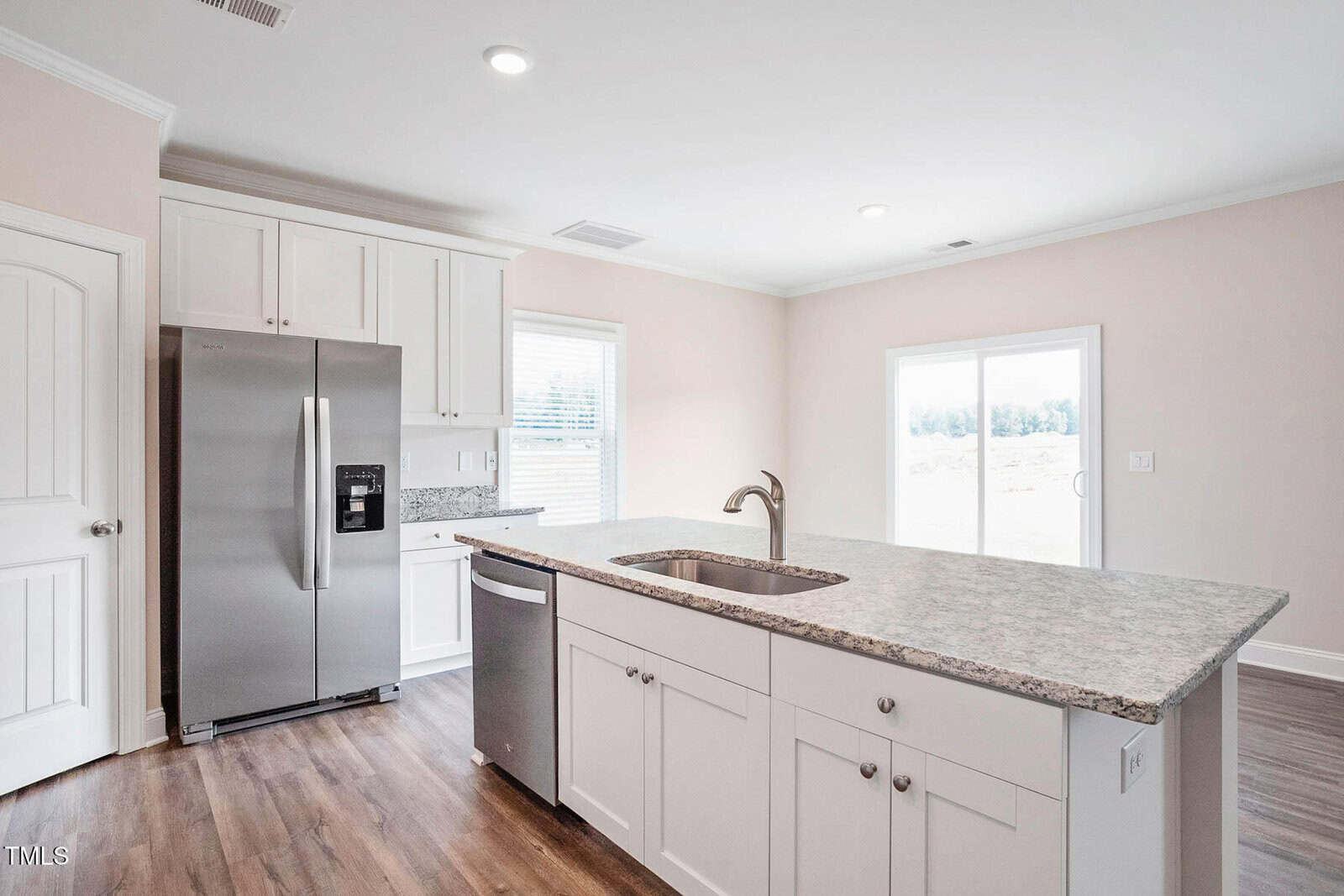 A modern kitchen with a granite countertop island, stainless steel fridge, and dishwasher. White cabinetry lines the walls, and a window and sliding glass door provide natural light. The floor is hardwood.