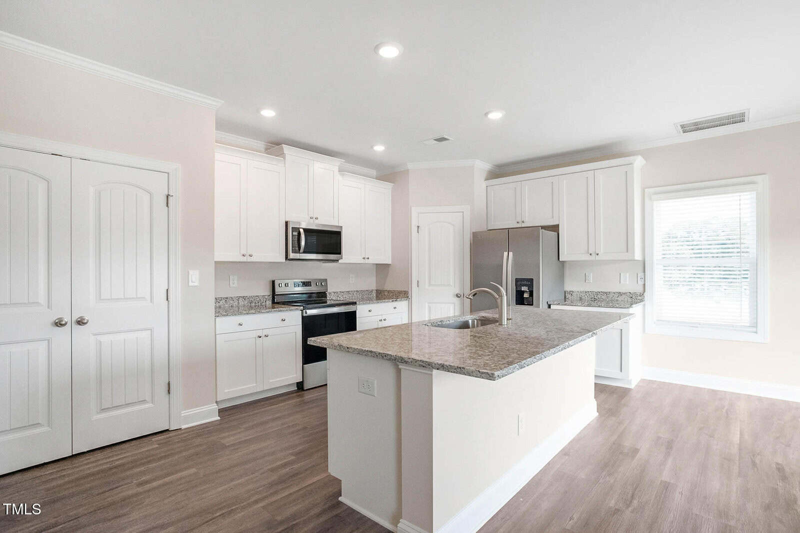 A modern kitchen with white cabinets, granite countertops, and stainless steel appliances, including a stove, microwave, and refrigerator. An island with a sink is in the center. Recessed lighting is on the ceiling, and a window on the right lets in natural light.