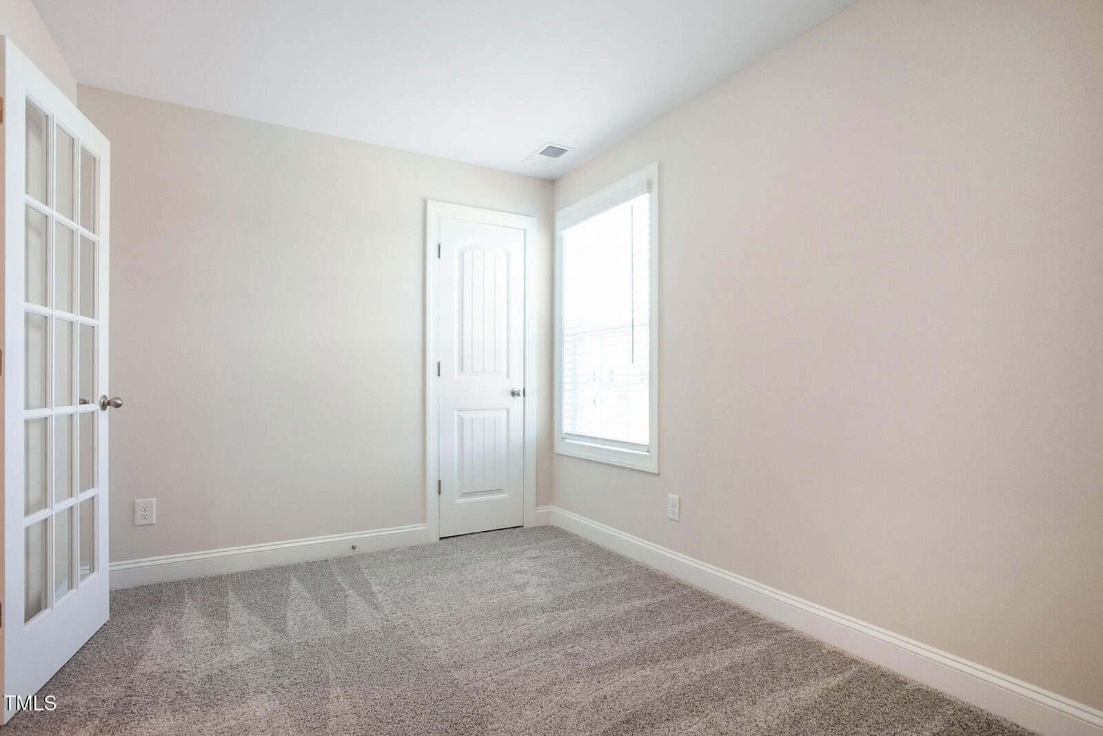 The image shows a small, empty room with beige walls and carpeted floor. There is a white door on one wall and a window with blinds on another. The room is well-lit with natural light.