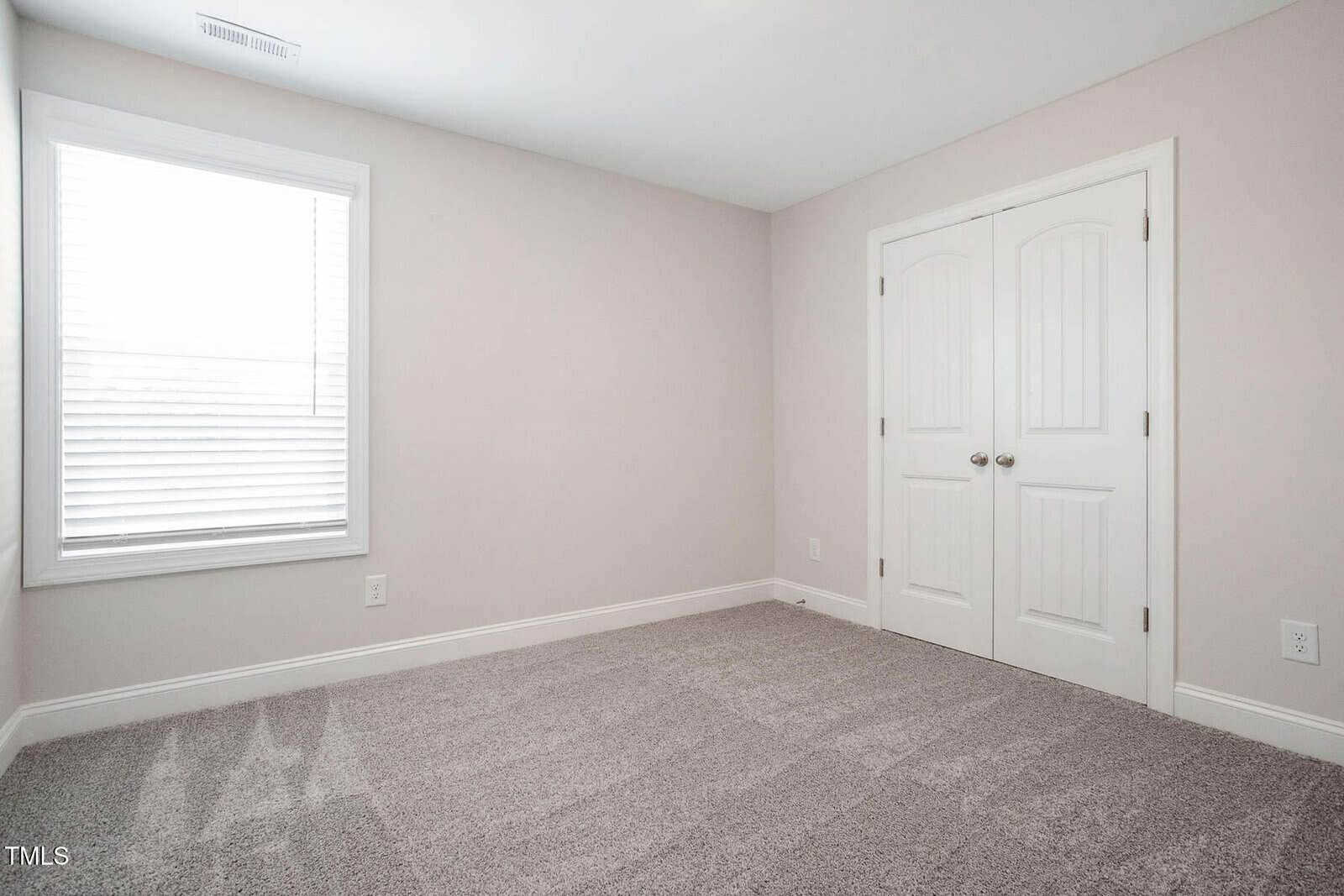 An empty room with beige walls, carpeted floor, and a window with closed blinds on the left. There is a double-door white closet on the right wall. The room appears to be well-lit by natural light.