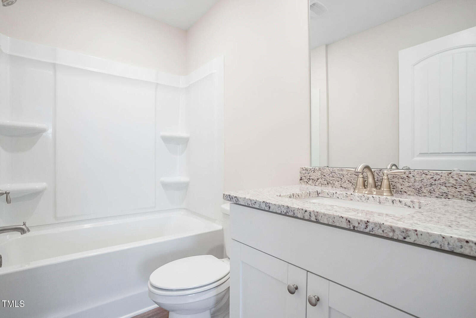A clean, modern bathroom featuring a white bathtub with a showerhead, a toilet, and a sink with a granite countertop. The wall is painted in a light color, and a large mirror is above the sink. The room is well-lit with natural and artificial light.