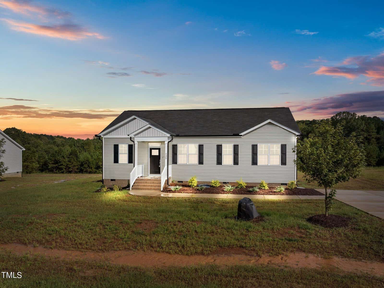 A single-story gray house with white trim and black shutters sits on a grassy lawn at sunset, with a front porch, landscaped garden, and a paved driveway. Trees and fields are visible in the background under a colorful sky.