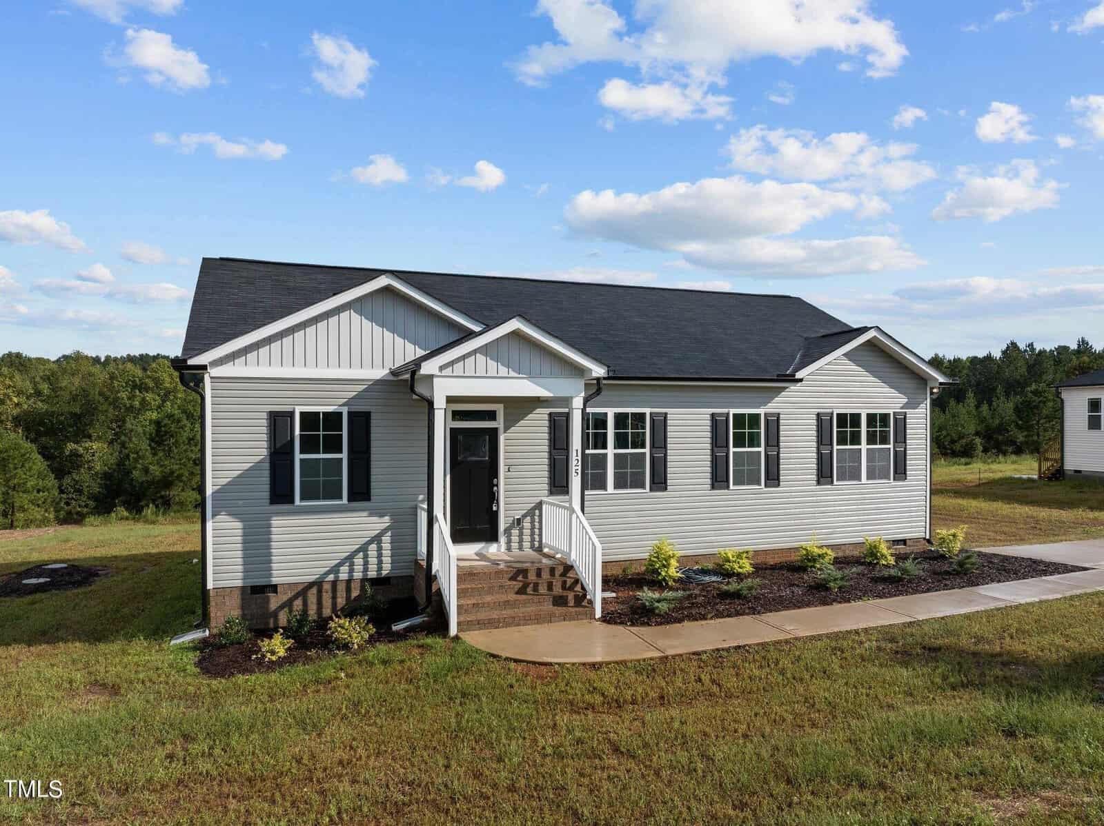 Single-story house with gray siding, black shutters, and a covered front porch. The house has a dark roof, a concrete walkway, and landscaped shrubs in front. Green trees and grass fill the background under a blue sky.